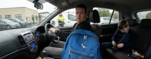 A blue child’s schoolbag in the foreground of a family car as a father checks the time and parents wait outside an Irish primary school. IMAGE TITLE: Seventeen Minutes Before School